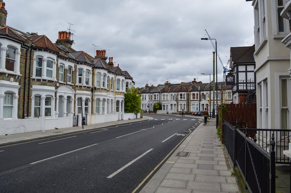 A narrow residential street in Clapham lined with brick terraced houses featuring multiple stories and chimneys, with some houses having small front gardens with green hedges and flowering plants. The