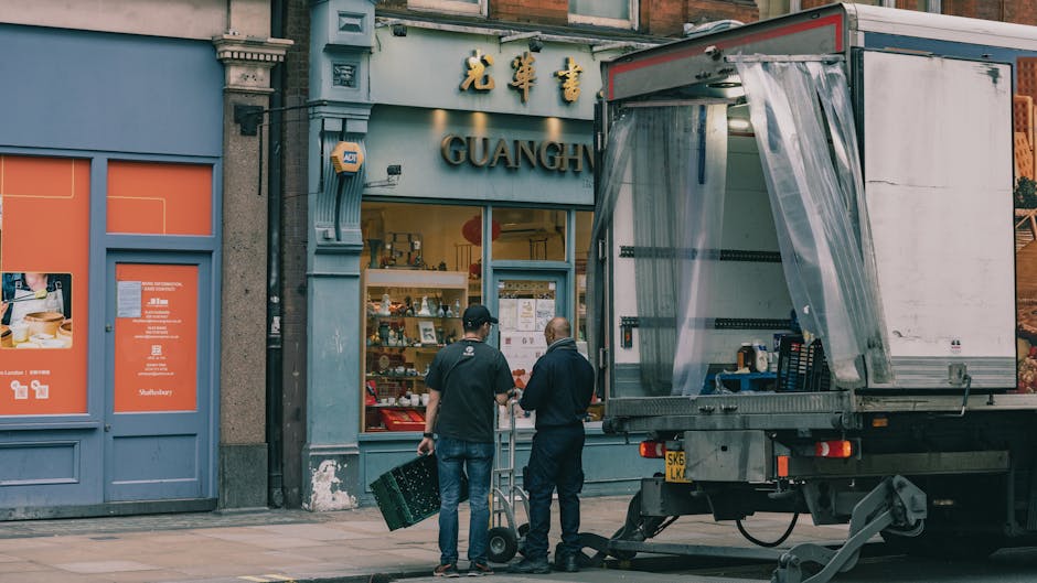 A person wearing a blue denim shirt and grey trousers is partially hidden behind a large cardboard box, holding a marker or pen in their right hand while marking the side of the box. The individual ha