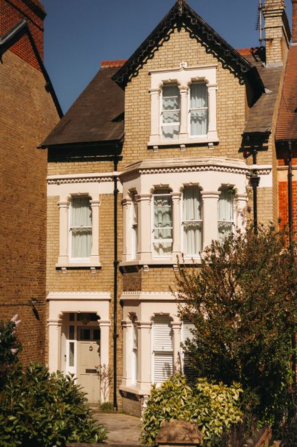 The front view of a three-storey Victorian-style house with a light brown brick facade, white decorative window surrounds, and a dark slate roof. The house features three tall windows on each floor, some with partially drawn curtains, and a white front door on the ground level. A small paved pathway leads to the entrance, which is partially obscured by green and yellow foliage in the front garden. The scene is bathed in natural daylight, highlighting the architectural details. This image is related to home relocation and furniture transport services offered by Man and a Van Clapham, illustrating a typical residential property suitable for house removals and moving logistics.