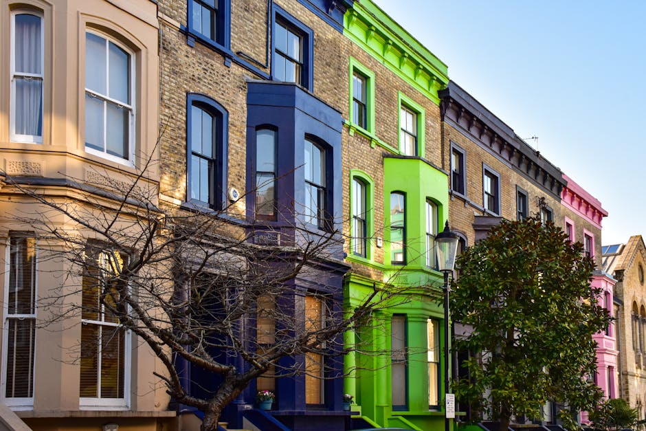 A row of four-story residential buildings with facades painted in pastel colors including beige, blue, green, and pink, situated along a street in Clapham. The buildings feature large sash windows, some with white frames, and decorative window sills. A leafless tree with multiple branches extends across the lower part of the image, partially obscuring the lower windows. A vintage-style black street lamp is positioned nearby, illuminating the scene. The photo is taken during daylight with a clear blue sky in the background. The vibrant green building has a small bay window on the second floor, while the other buildings display characteristic Victorian architectural details. The scene suggests an urban environment suitable for home relocation or furniture transport, with the colorful facades adding visual interest for a house removals guide related to flats and houses in Clapham, as seen on manandavanclapham.co.uk.