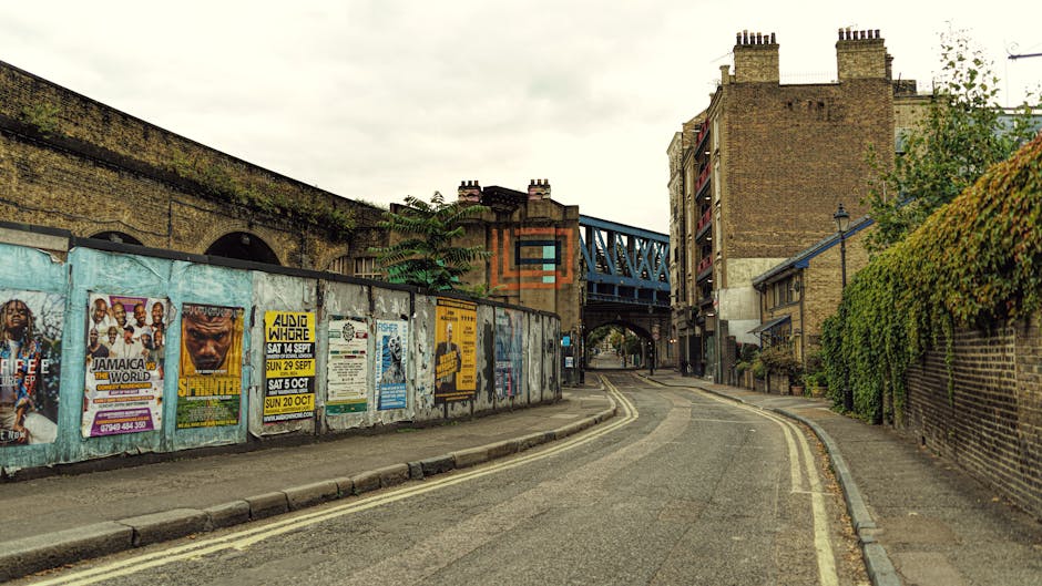 A narrow residential street in Clapham with an upward curve and a slight incline, featuring a row of brick houses on the right side with green foliage growing along the wall. On the left, a metal barr
