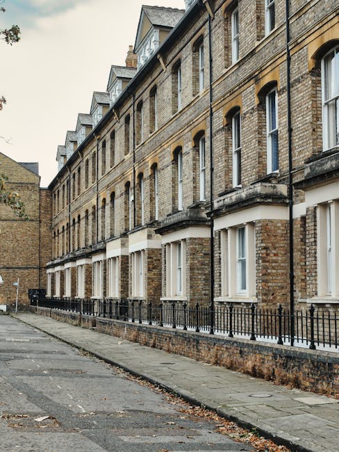 A narrow residential street in Clapham with an upward curve and a slight incline, featuring a row of brick houses on the right side with green foliage growing along the wall. On the left, a metal barr