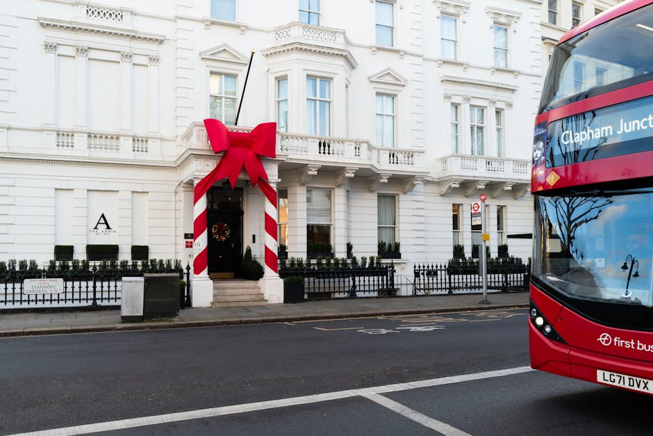 A white, multi-storey building on a city street, decorated for the holiday season with a large red bow and candy stripe-patterned ribbon framing the entrance. The building's facade features tall windows, small balconies, and decorative architectural details. Black metal fencing runs along the front, dividing the sidewalk from the building. In the foreground, a red double-decker bus with the route label 'Clapham Junction' is partially visible on the right side, indicating a busy urban environment. The street has a marked lane and is generally clear of traffic, with a few street signs and a small trash bin nearby. The scene captures a typical day in Clapham, with a context suitable for house relocation or moving services provided by Man and a Van Clapham, which may be involved in home relocation, furniture transport, or packing and moving logistics.
