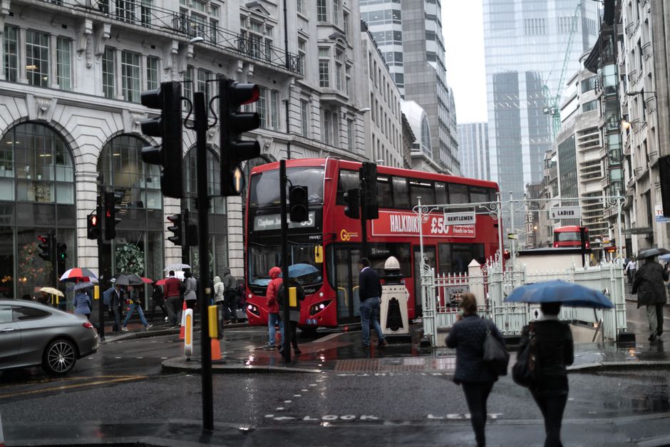 A busy city street view in Clapham Junction with two red double-decker buses navigating through traffic and pedestrians crossing the wet road holding umbrellas. The background features tall modern office buildings with glass facades, and older white stone structures, reflecting typical urban architecture. In the foreground, there are black and yellow traffic signals, and several people dressed in coats and umbrellas, walking across the crossing and waiting on the sidewalk. A grey car is visible on the left side of the image, and a white temporary barrier is positioned near the bus stop area. This scene captures a rainy day in central London, illustrating urban transport and pedestrian activity consistent with house removals and moving logistics in the area, with some individuals approaching the buses likely to be involved in home relocation or furniture transport supported by local removals services such as Man and a Van Clapham.